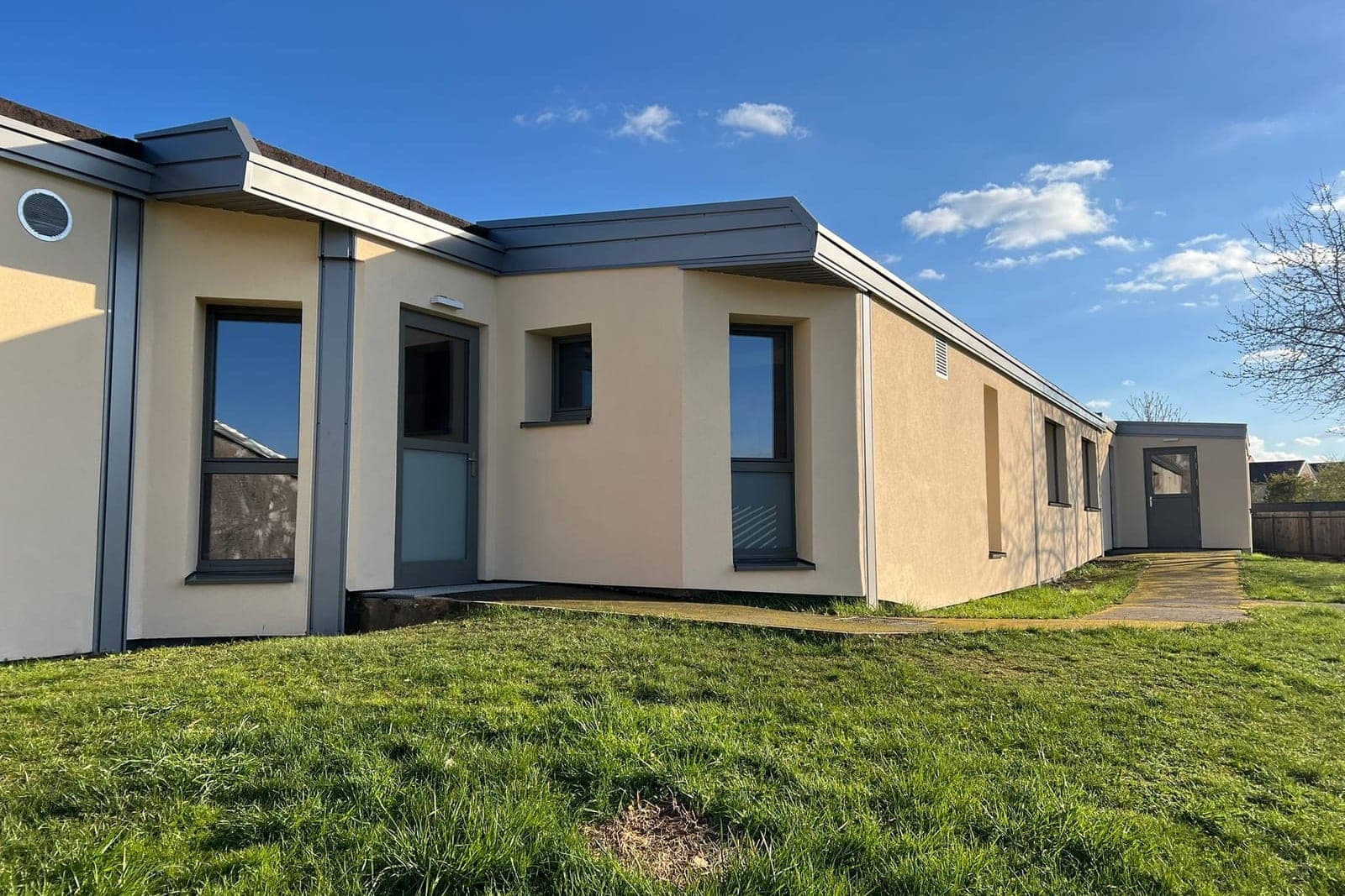 École de plain-pied : enduit crème, habillages de toiture gris, grandes baies vitrées et pelouse au premier plan, sous un ciel bleu.