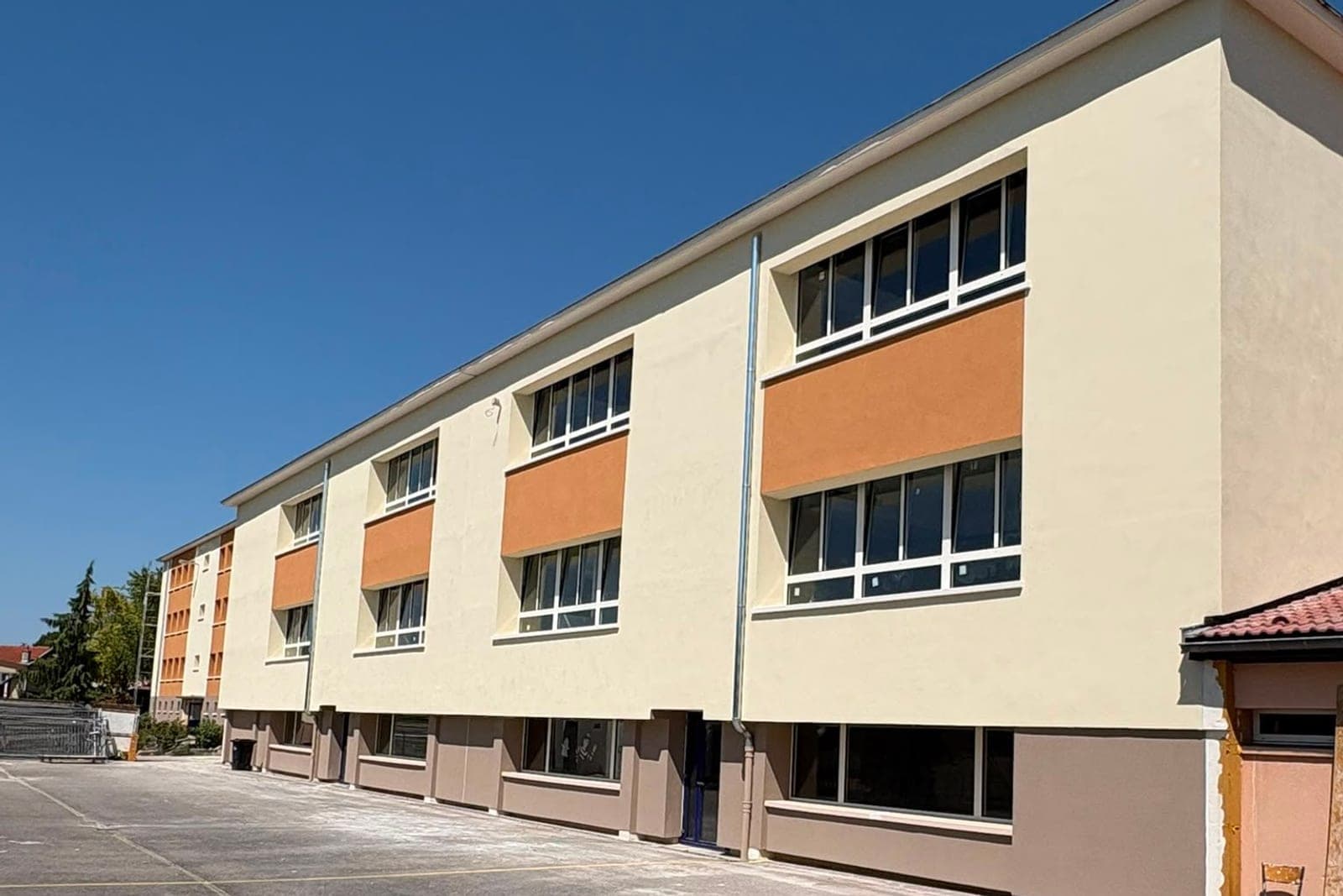 Bâtiment scolaire à trois niveaux sur sa cour : enduit crème, bandeaux orangés entre les fenêtres, soubassement taupe, sous un ciel bleu.