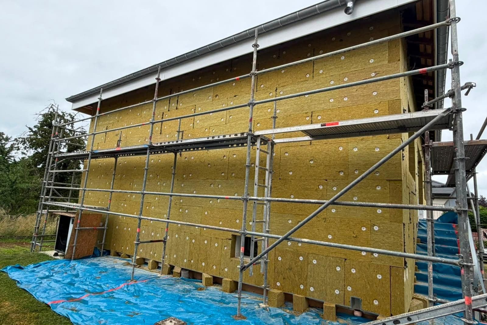 Pignon d'une maison sous échafaudage, entièrement couvert de panneaux isolants jaunes fixés par chevilles, bâche bleue de protection au sol.