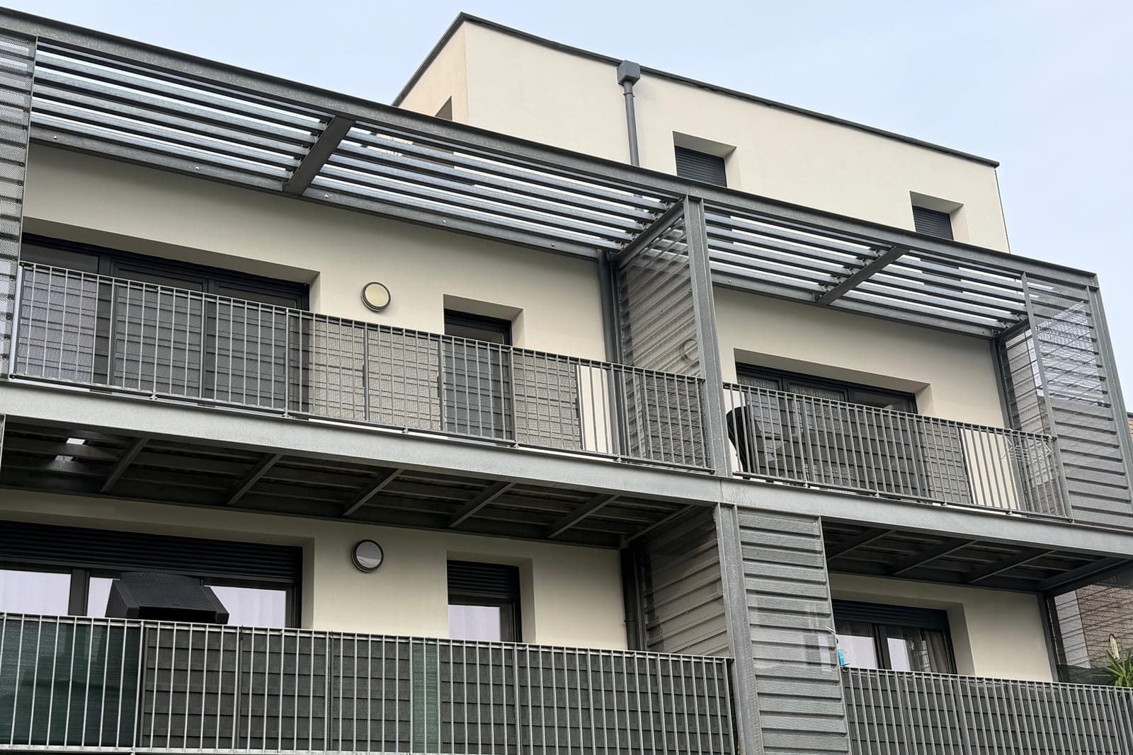 Façade d'une résidence récente vue d'en bas : enduit clair, balcons à garde-corps métalliques et habillages en bardage gris à lames horizontales, pergola à lames au dernier étage.